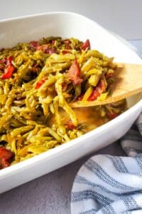 A wooden spoon lifts a serving of Green Beans with Bacon and Onion, mixed with tomato, from a white baking dish.