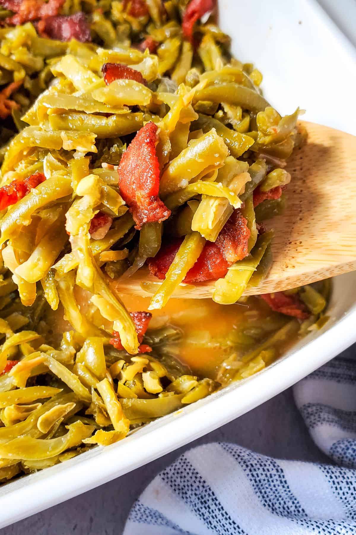 A wooden spoon holds a serving of Green Beans with Bacon and Onion, featuring tender green beans mixed with savory bacon pieces in a white dish.