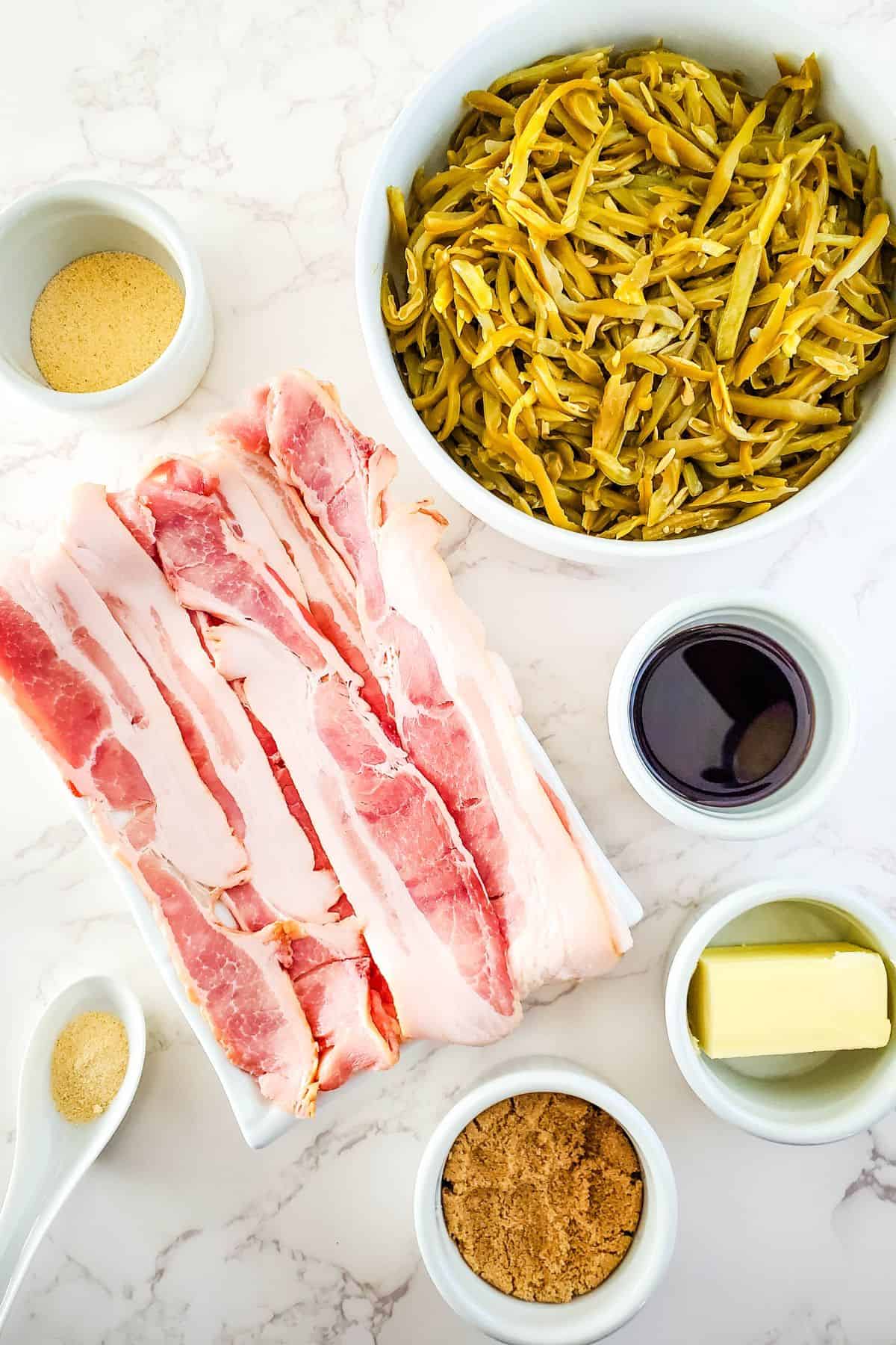 Ingredients, raw bacon strips, canned green beans, brown sugar, butter, soy sauce, garlic powder, and onion powder—arranged in bowls on a white marble surface.