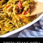 A wooden spoon scoops Green Beans with Bacon and Onion from a white dish, sitting on a striped cloth.