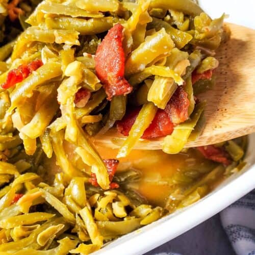 A wooden spoon holds a serving of Green Beans with Bacon and Onion, perfectly cooked and mixed in a white dish.