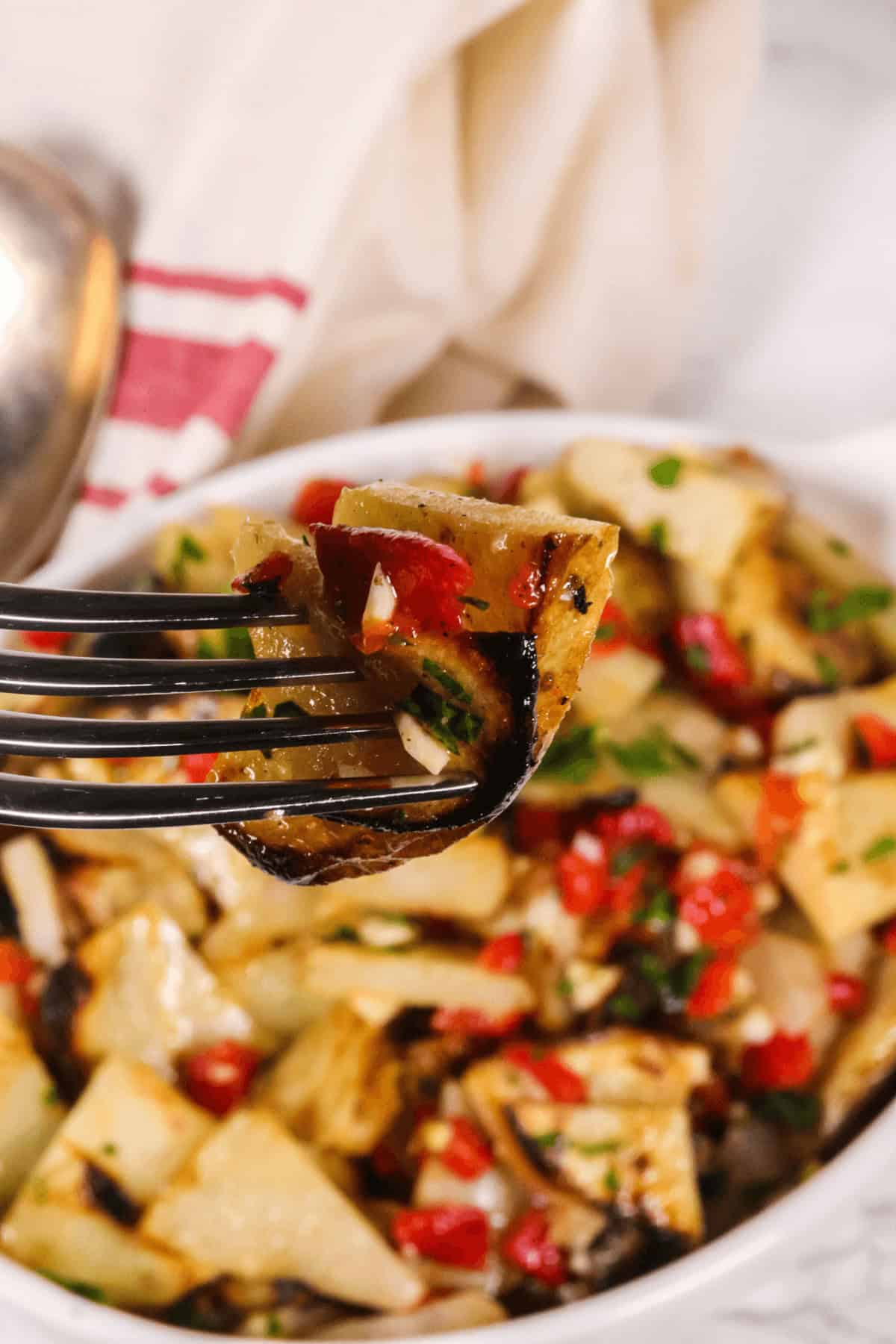 A close-up of a fork holding a piece of roasted potato with herbs and diced red peppers. In the background, a bowl of warm potato salad is artfully blurred. A red-striped towel is partially visible.