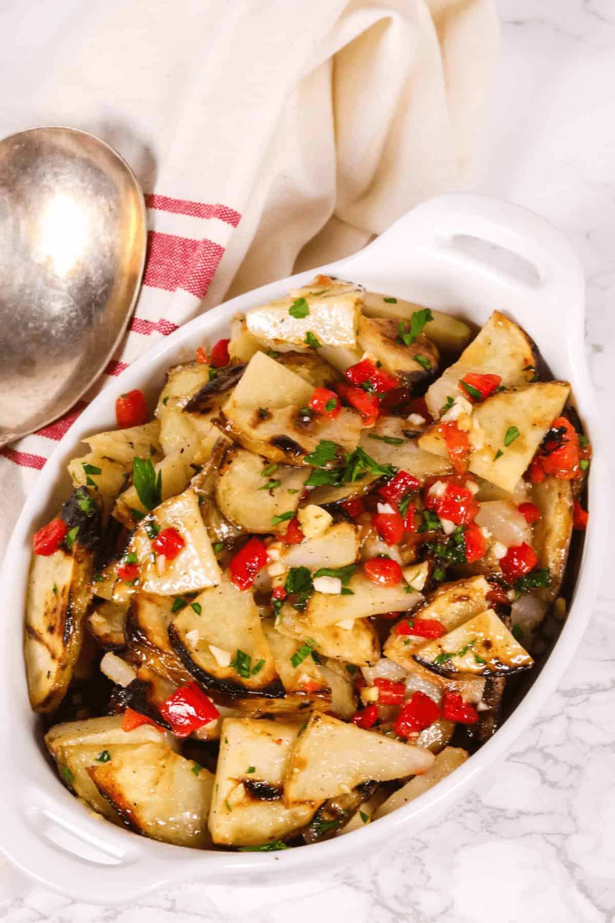 A warm potato salad fills a white dish, featuring grilled potato pieces, chopped red peppers, and herbs. It sits on a marble surface next to a spoon and a white and red-striped cloth.