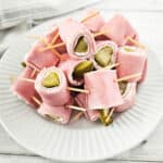 A plate of pickle roll-ups with ham, secured by toothpicks, sits on a pristine white dish against a striped cloth backdrop.