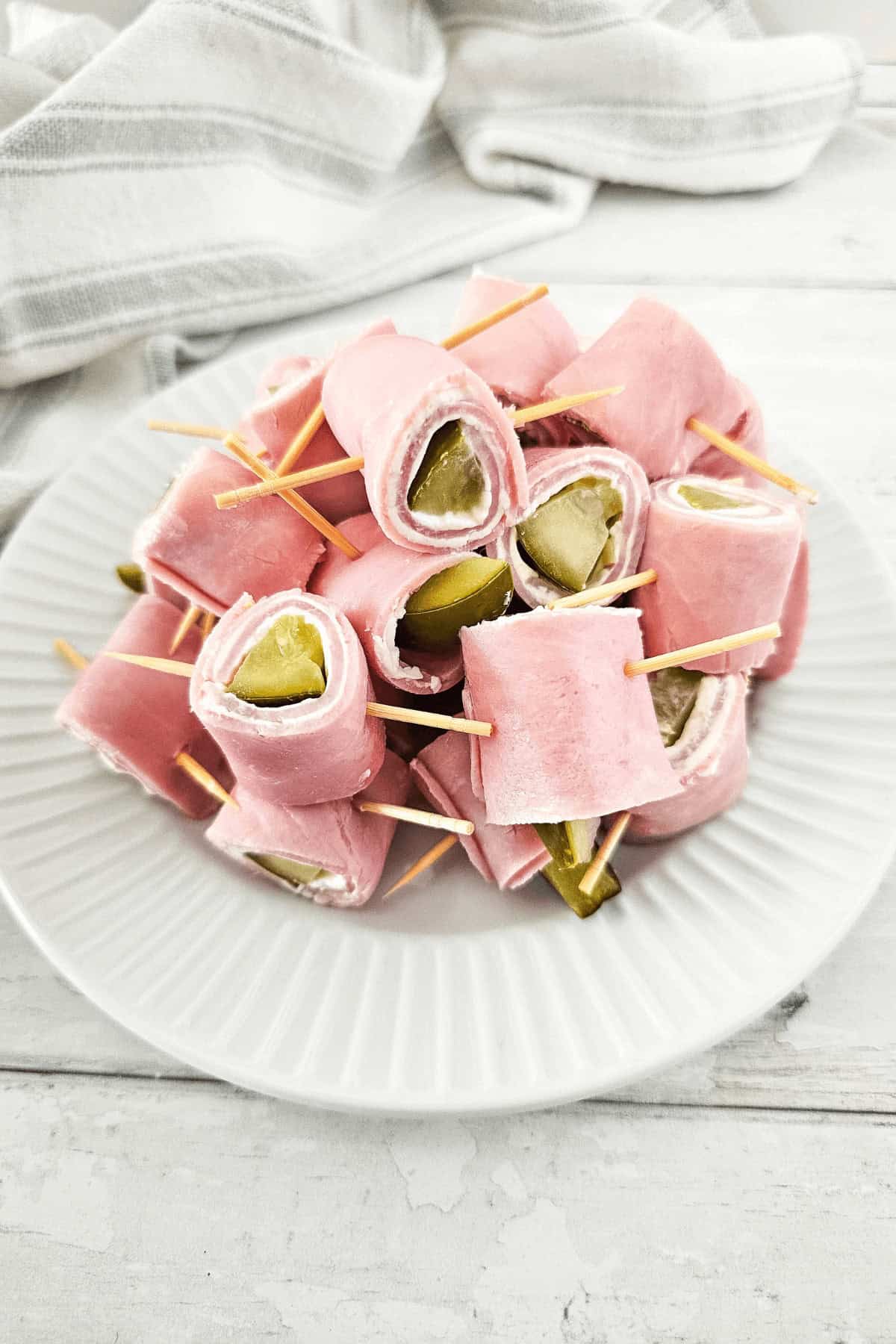 A plate of pickle roll-ups with ham, secured by toothpicks, sits on a pristine white dish against a striped cloth backdrop.