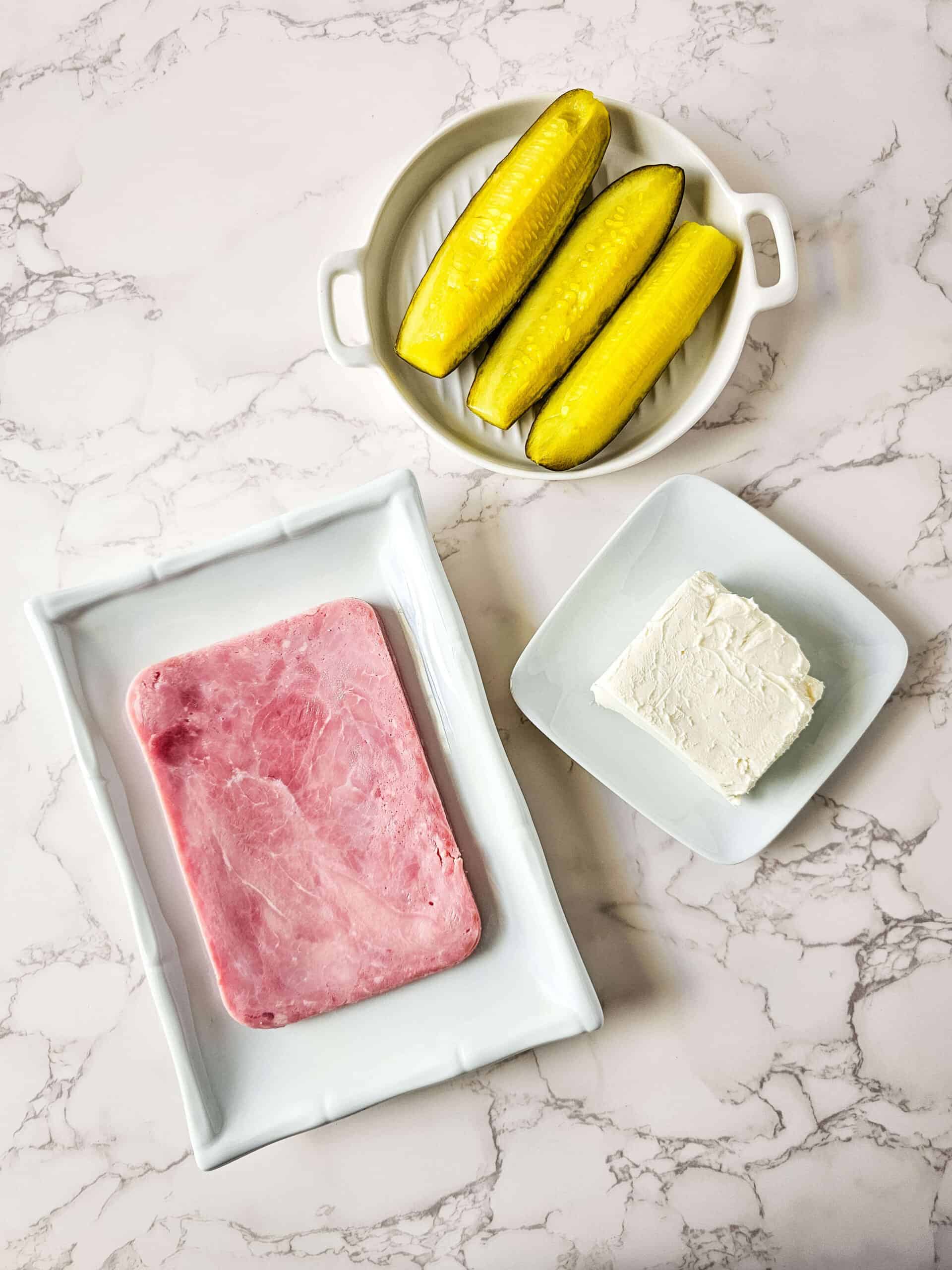 Sliced ham on a rectangular plate, a block of cream cheese on a square dish, and three pickles in a round dish create the perfect setup for making pickle roll ups, all elegantly displayed on a marble surface.