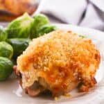 A Hellman’s Crusted Parmesan Chicken thigh with a golden, crispy crust is served on a white plate alongside steamed Brussels sprouts.