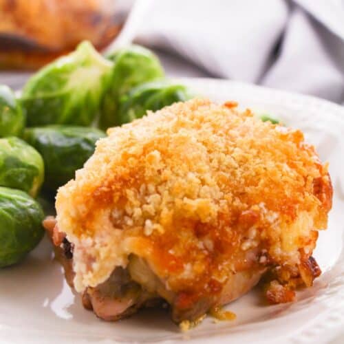 A Hellman’s Crusted Parmesan Chicken thigh with a golden, crispy crust is served on a white plate alongside steamed Brussels sprouts.