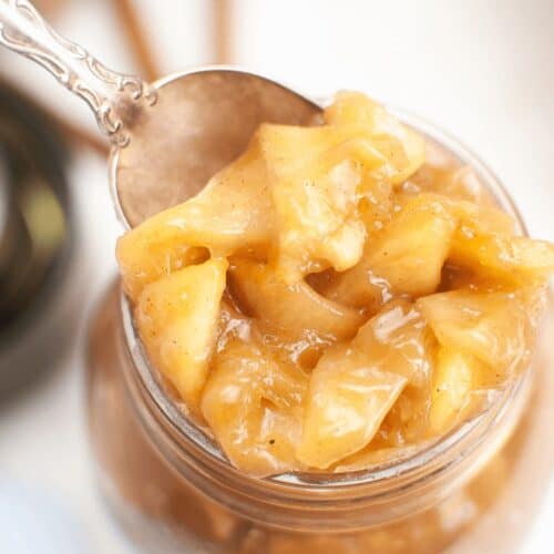 A close-up of a spoonful of chunky homemade apple pie filling being lifted from a glass jar.