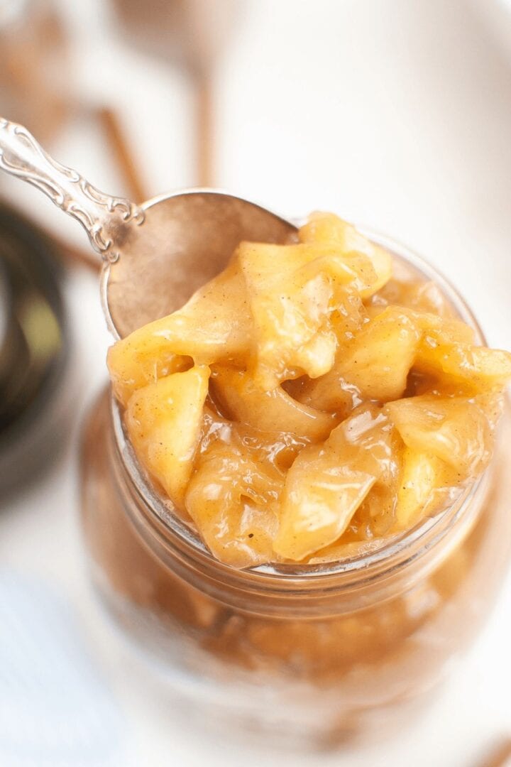 A close-up of a spoonful of chunky homemade apple pie filling being lifted from a glass jar.