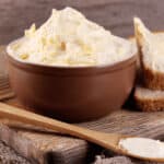 A bowl of homemade butter sits on a wooden board next to a buttered slice of bread and a wooden knife.