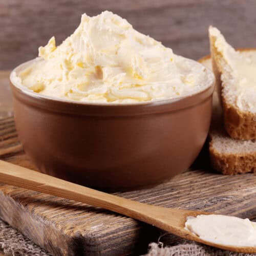 A bowl of homemade butter sits on a wooden board next to a buttered slice of bread and a wooden knife.