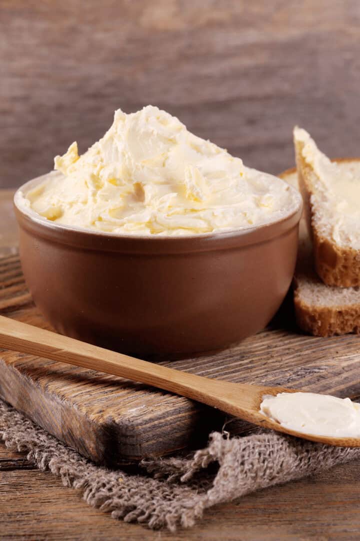 A bowl of homemade butter sits on a wooden board next to a buttered slice of bread and a wooden knife.