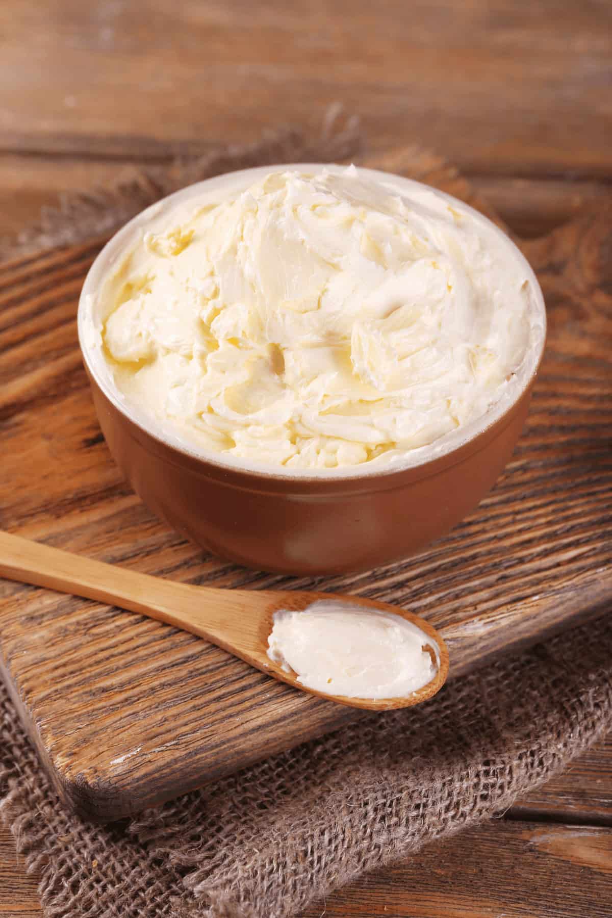 A wooden bowl filled with homemade whipped butter sits on a wooden board, accompanied by a small wooden spoon with a dab of creamy butter on it.