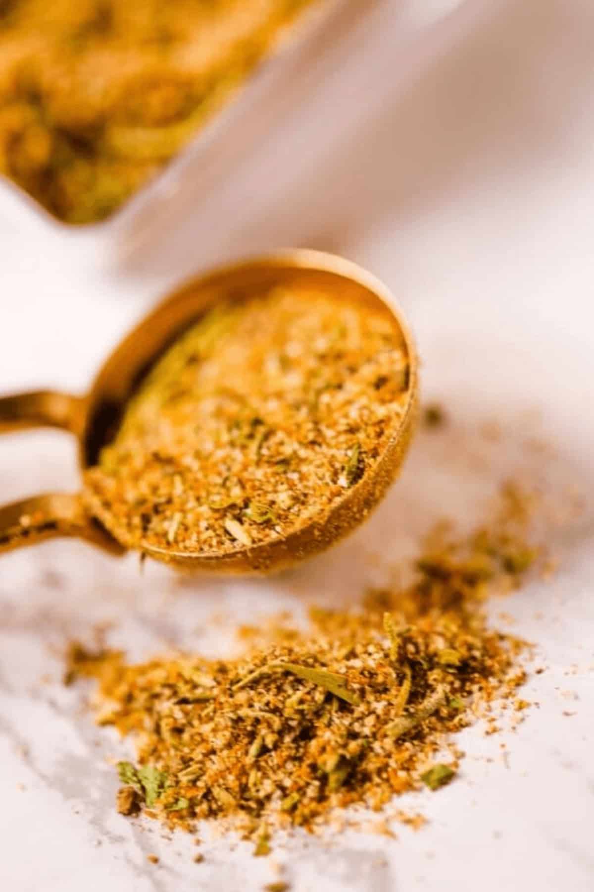 A measuring spoon filled with a rich blend of homemade chili seasoning rests on a marble surface, surrounded by a scattering of vibrant spices and herbs.