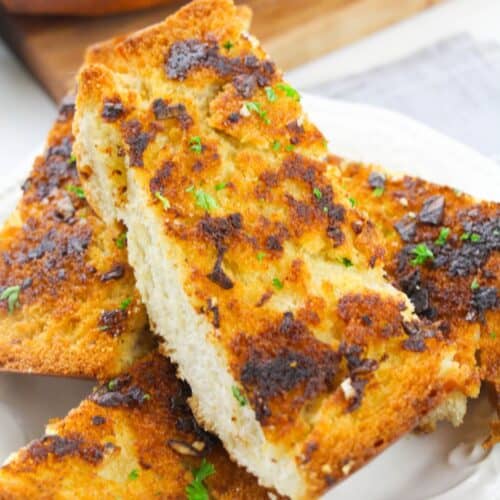 Three slices of homemade garlic bread with a golden, crispy crust are stacked on a white plate, garnished with chopped herbs.