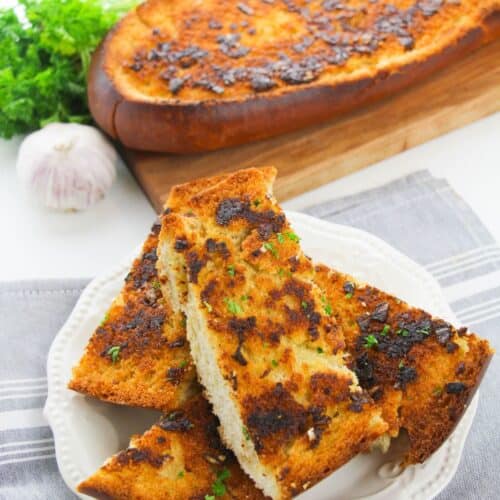 Slices of toasted Homemade Garlic Bread are served on a white plate, with a larger loaf and fresh herbs in the background.
