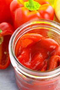 A glass jar filled with vegetables in liquid, surrounded by whole and sliced red bell peppers.