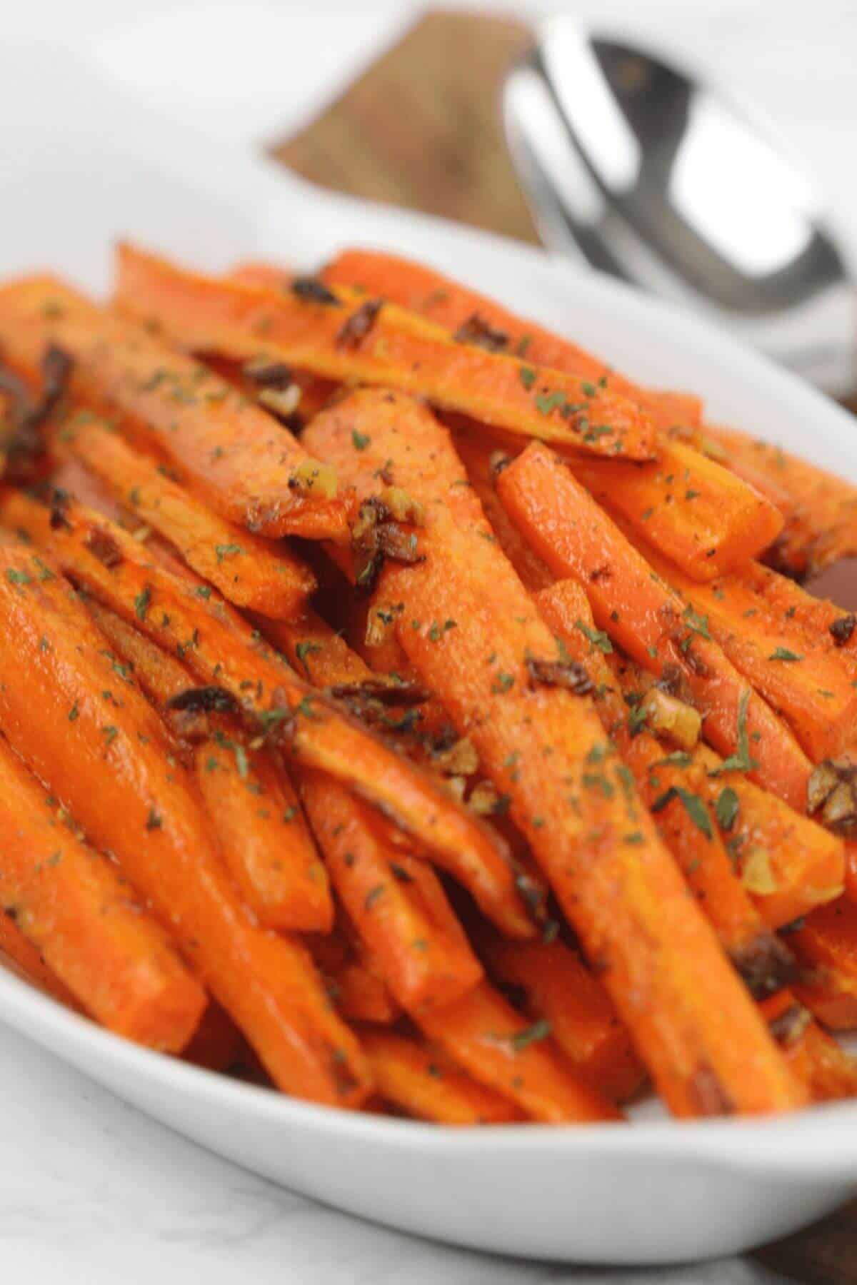A white dish filled with roasted baby carrots cut into sticks, garnished with chopped herbs and small pieces of garlic.