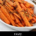 A white dish filled with Roasted Baby Carrots, garnished with herbs and bits of garlic, with serving utensils in the background.
