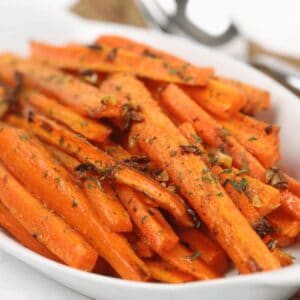 A white dish filled with roasted baby carrots, garnished with fresh herbs and small pieces of garlic.