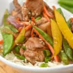 A bowl of rice topped with honey sesame chicken pieces, snow peas, carrots, yellow bell peppers, and red onions.
