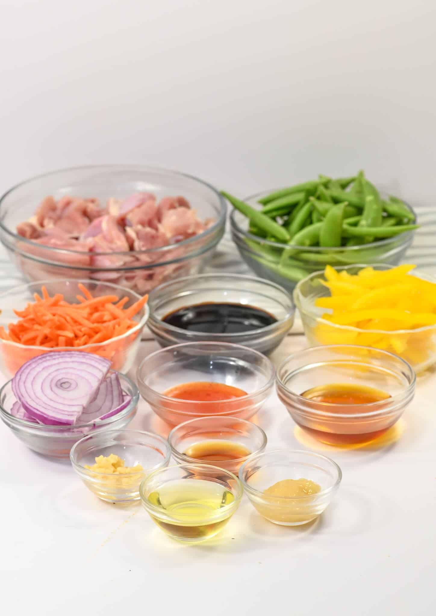 A variety of ingredients in bowls, including sliced chicken, snap peas, sliced onion, shredded carrots, and yellow peppers; a vibrant display reminiscent of Honey Sesame Chicken with several sauces and oils arranged on a white surface.