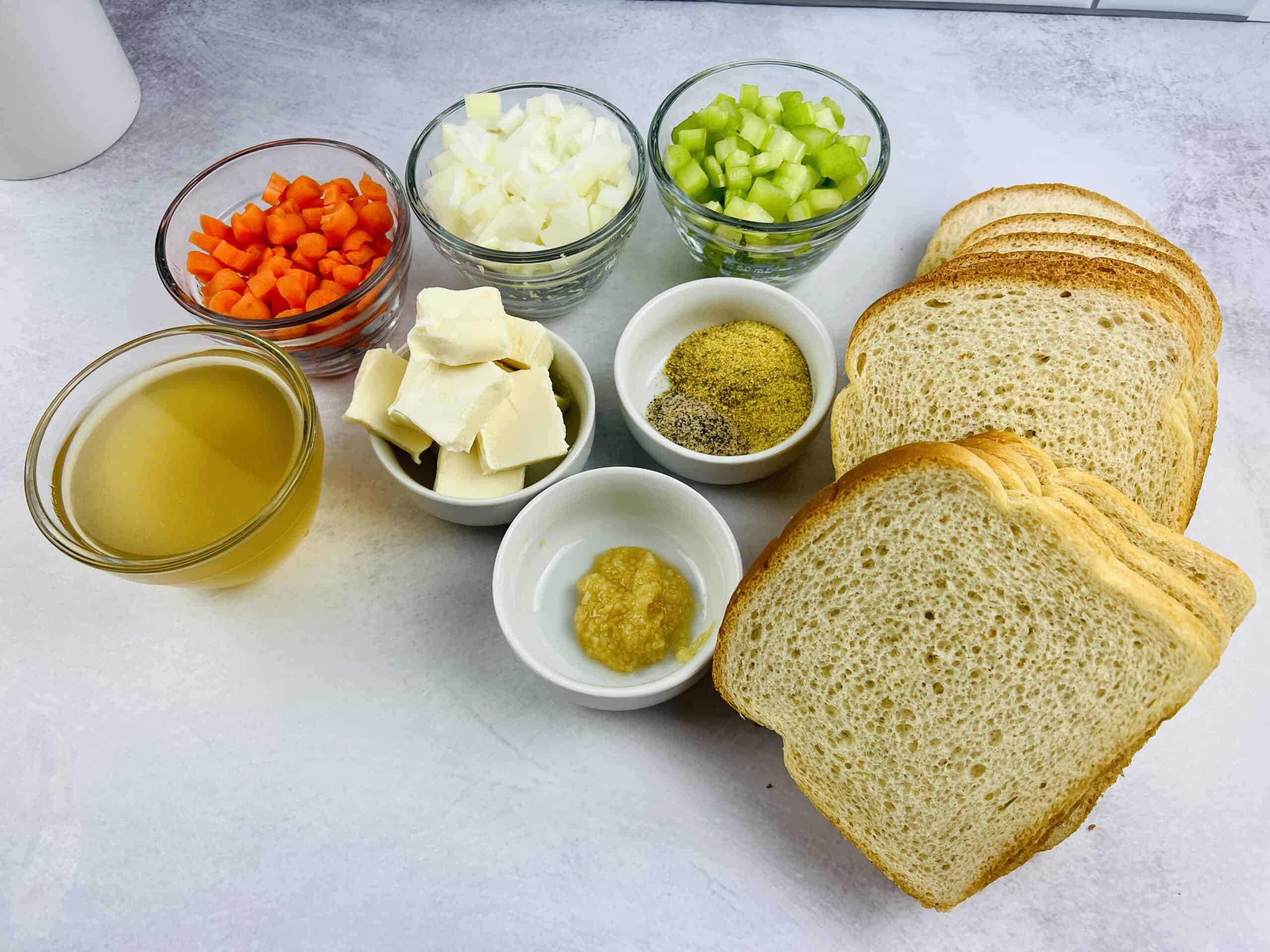 Sliced white bread, chopped carrots, onions, celery, small bowls of butter, broth, garlic paste, black pepper, and herbs arranged on a white surface—perfect for making a classic Stove Top Stuffing Recipe.