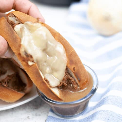A hand holds a meal with melted cheese, dipping it into a small bowl of sauce, creating the perfect Instant Pot French Dip moment. A striped blue and white cloth rests on the table, adding to the cozy atmosphere.