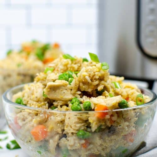 A clear glass bowl filled with Instant Pot chicken fried rice, featuring peas, carrots, and green onions, sits in the foreground with a kitchen appliance in the background.
