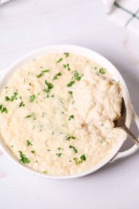 A bowl of creamy side dish topped with chopped parsley, with a spoon elegantly lifting a portion from the side.