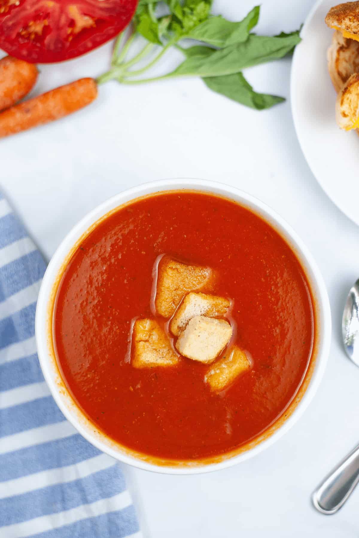 A bowl of Instant Pot tomato soup topped with croutons sits invitingly. Nearby, a striped cloth cradles a spoon, with sliced tomatoes, fresh carrots, fragrant herbs, and bread slices completing the cozy scene.