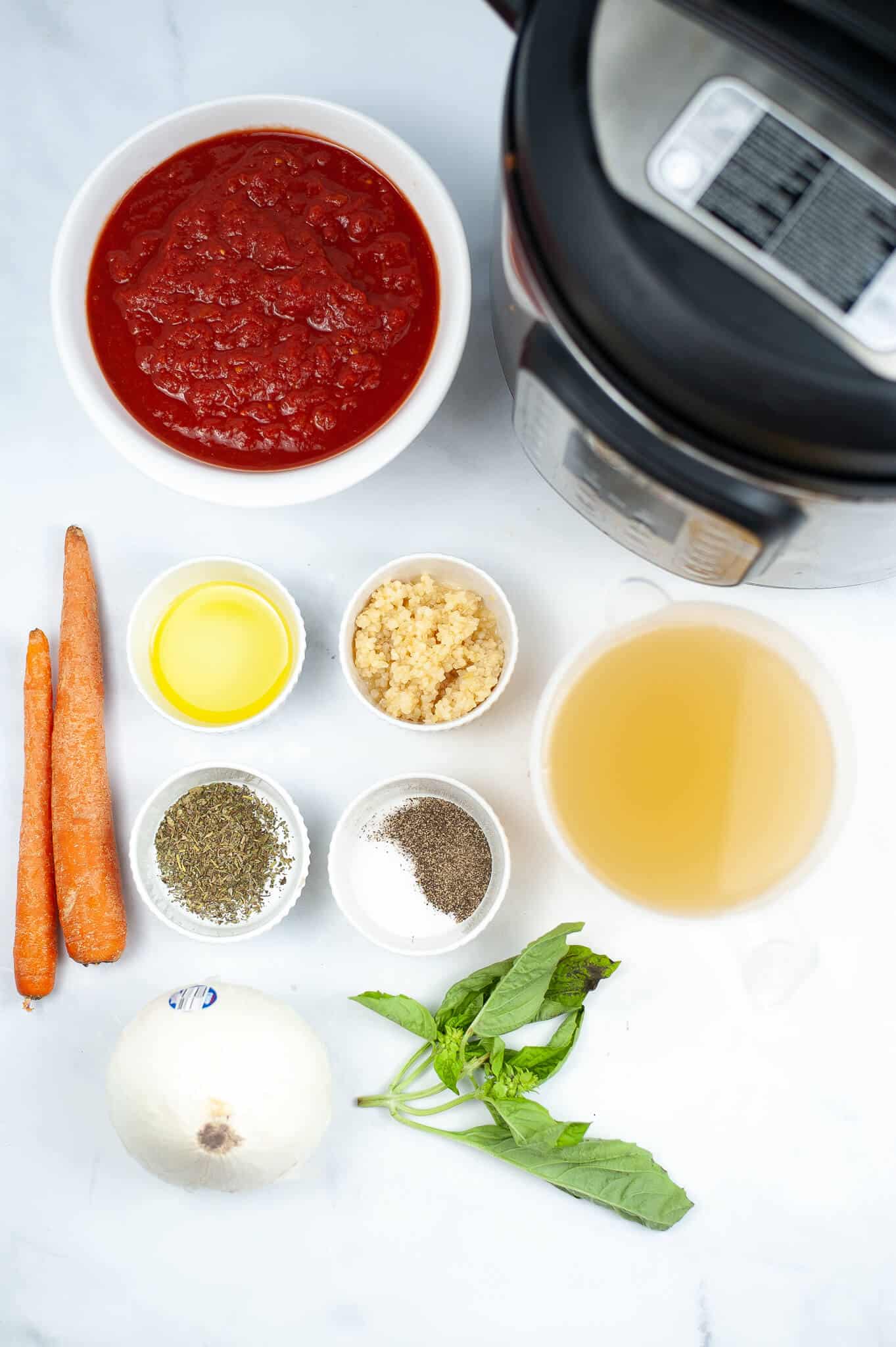 Ingredients for a delicious Instant Pot Tomato Soup are arranged neatly: canned tomatoes, carrots, olive oil, minced garlic, broth, herbs, spices, onion, and fresh basil.