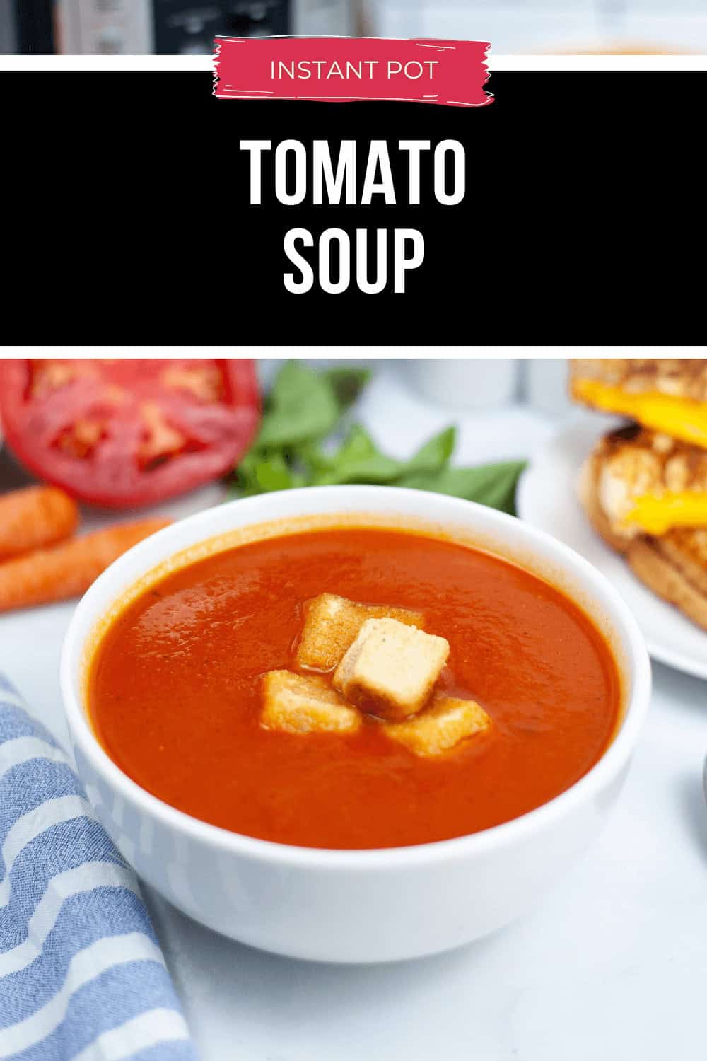 A bowl of Instant Pot tomato soup is topped with crispy croutons and surrounded by fresh carrots, a sliced tomato, and a hearty sandwich.