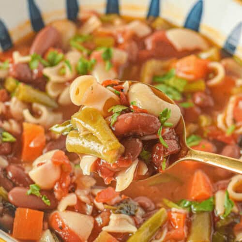 A bowl of Italian minestrone soup brimming with pasta, kidney beans, carrots, and green beans. A spoon is lifting a delicious portion of the classic soup.