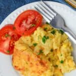 A serving of Jiffy Cornbread Casserole garnished with chopped parsley, accompanied by two slices of fresh tomato on a white plate with a fork.