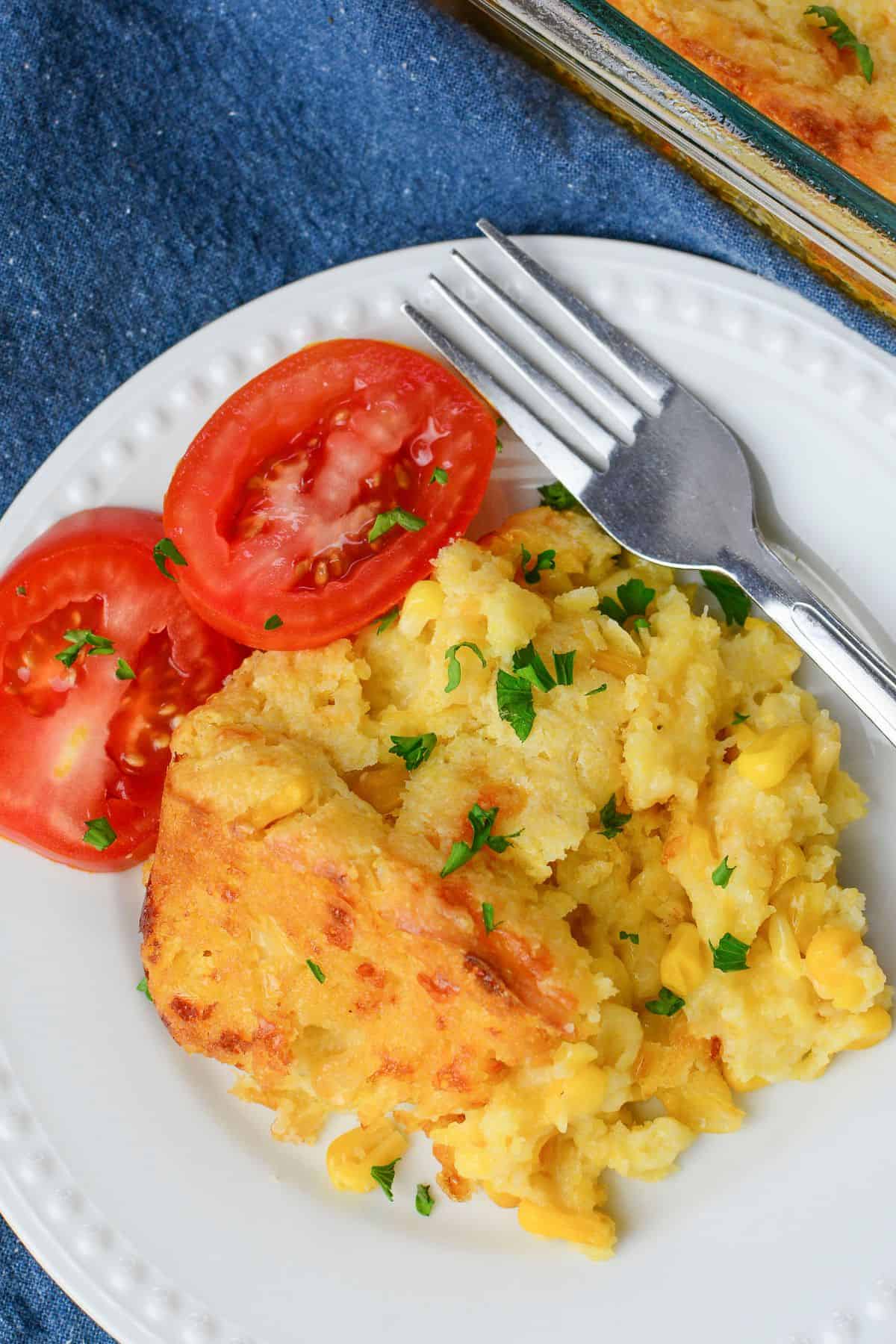 A serving of Jiffy Cornbread Casserole garnished with chopped parsley, accompanied by two slices of fresh tomato on a white plate with a fork.