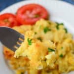 A fork holds a bite of cheesy Jiffy Cornbread Casserole garnished with parsley, with sliced tomatoes and more casserole on a white plate in the background.