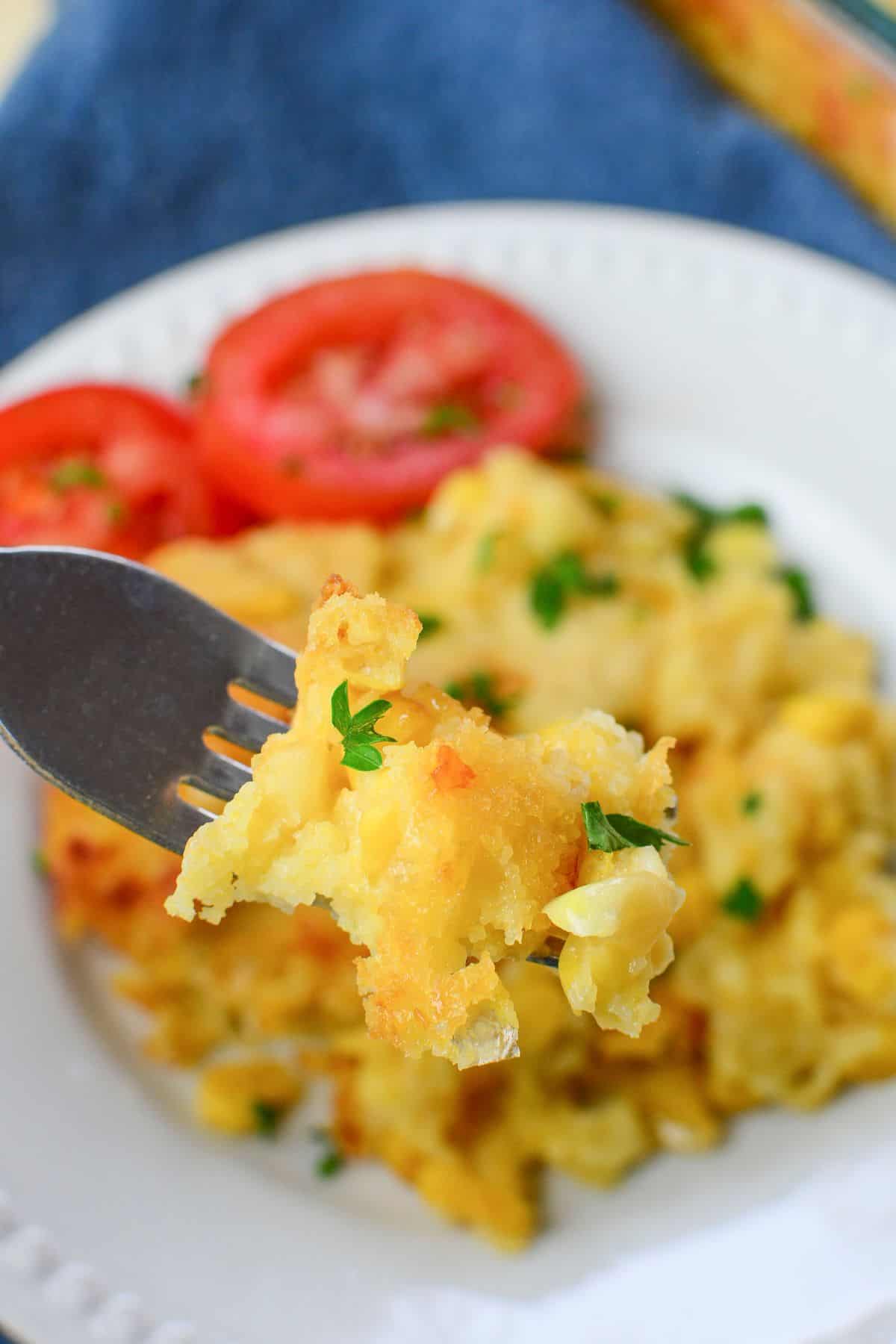 A fork holds a bite of cheesy Jiffy Cornbread Casserole garnished with parsley, with sliced tomatoes and more casserole on a white plate in the background.