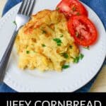A serving of Jiffy Cornbread Casserole garnished with parsley, served alongside sliced tomatoes on a white plate with a fork.