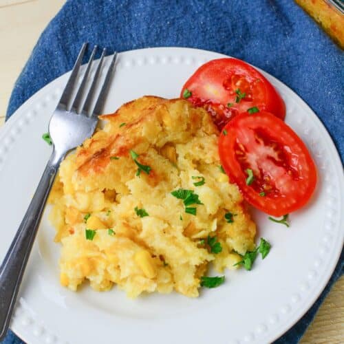A serving of cheesy Jiffy Cornbread Casserole with chopped parsley and sliced tomatoes on a white plate, placed on a blue cloth with a fork.
