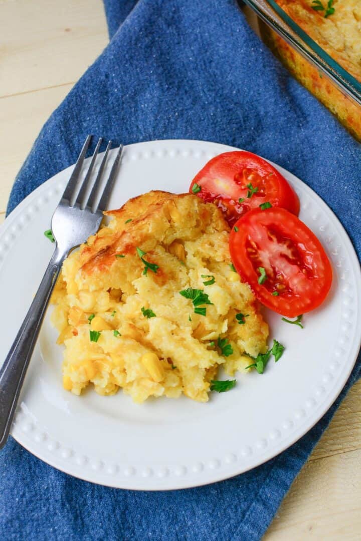 A serving of cheesy Jiffy Cornbread Casserole with chopped parsley and sliced tomatoes on a white plate, placed on a blue cloth with a fork.