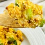 A wooden fork holds a serving of Jiffy Cornbread Dressing, with visible chunks of celery and onions, above a white plate.