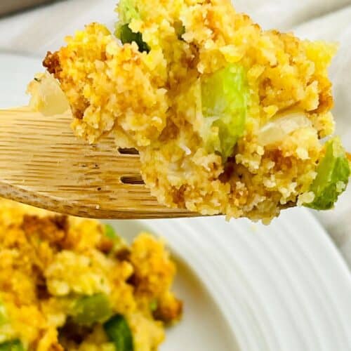 A wooden fork holds a serving of Jiffy Cornbread Dressing, with visible chunks of celery and onions, above a white plate.