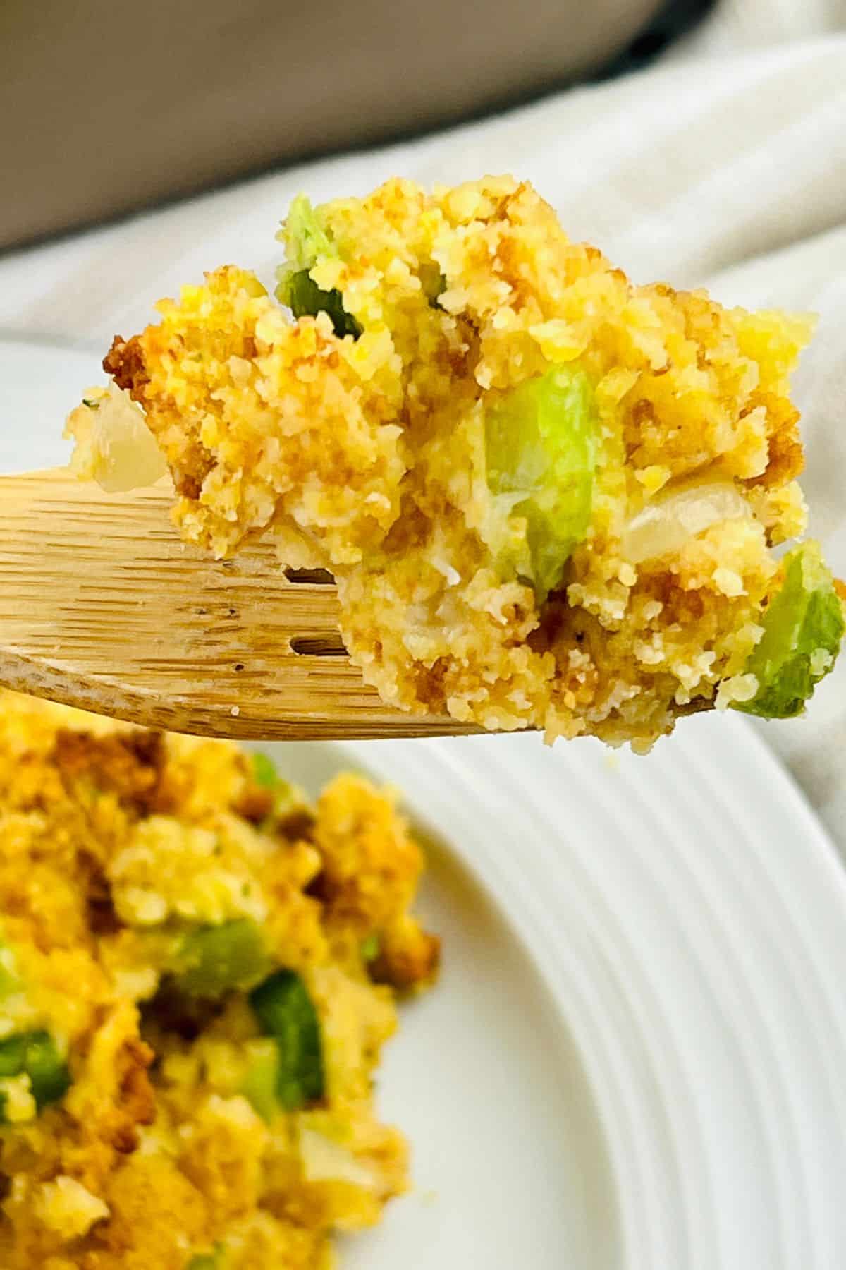 A wooden fork holds a serving of Jiffy Cornbread Dressing, with visible chunks of celery and onions, above a white plate.