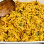 A close-up of a baking dish filled with Jiffy Cornbread Dressing, mixed with vegetables, and a wooden spoon resting on the side.