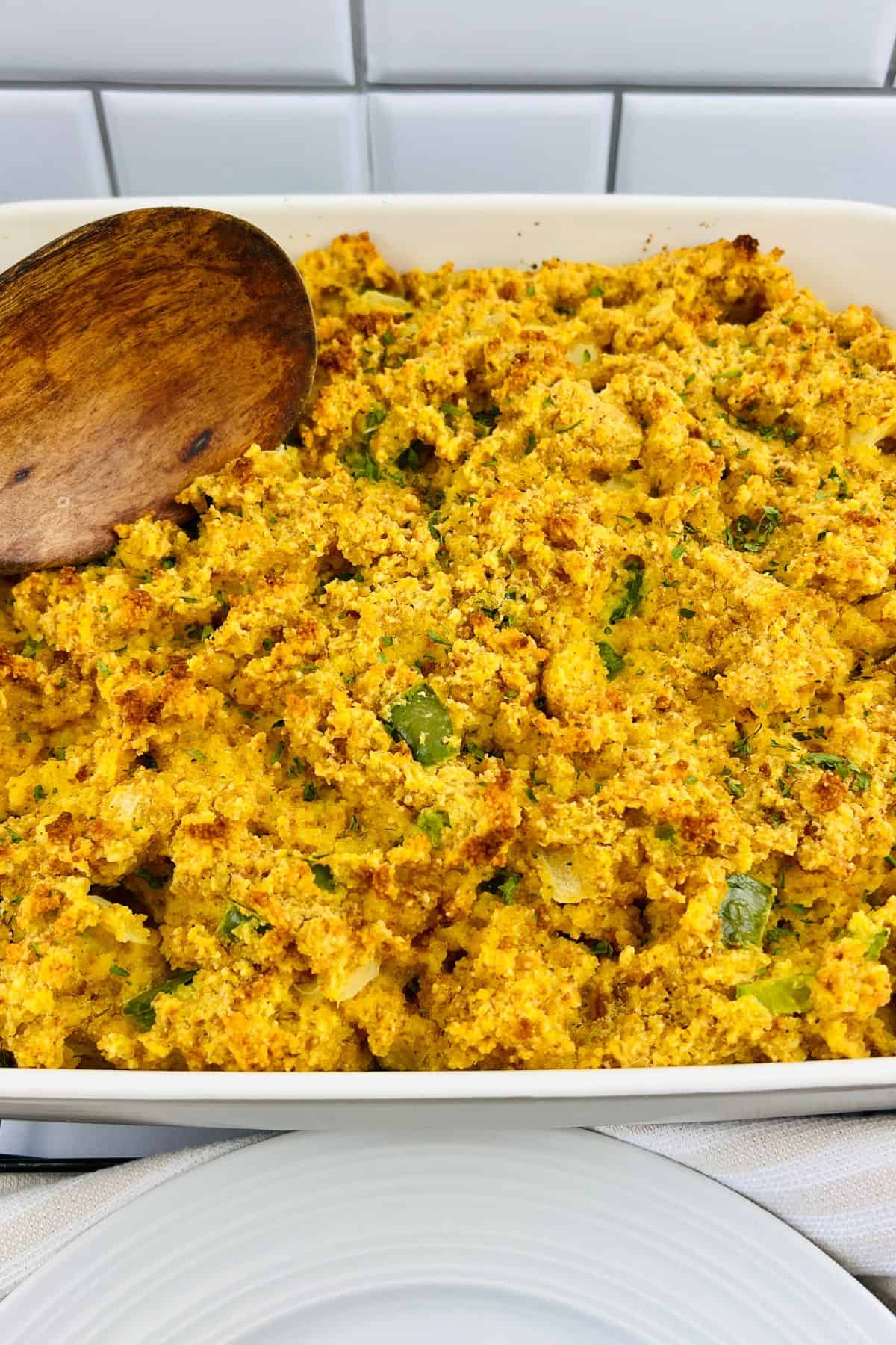 A close-up of a baking dish filled with Jiffy Cornbread Dressing, mixed with vegetables, and a wooden spoon resting on the side.