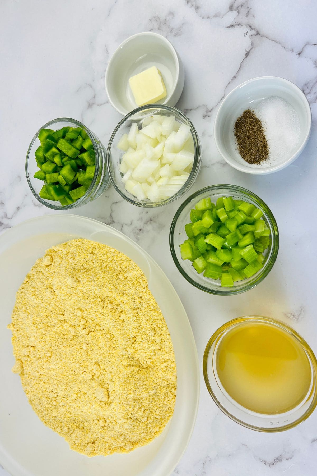 Bread, chopped green bell pepper, celery, onion, a pat of butter, broth, and bowls of salt and pepper are arranged on a marble surface.