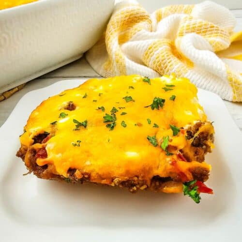 A slice of cheesy John Wayne Casserole with ground meat and vegetables sits on a white plate, with a baking dish and a yellow-and-white towel in the background.