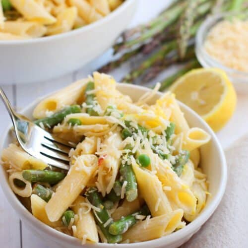 A bowl of Lemon Asparagus Pasta features penne mixed with asparagus and topped with grated cheese, accompanied by a fork, lemon, and fresh asparagus spears in the background.